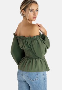 Olive green off-the-shoulder top with ruffled neckline, smocked waist, and long puff sleeves, paired with light blue jeans.