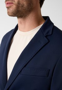 Marineblazer met een gladde textuur, gestikte revers en een minimalistische borstzak, gecombineerd met een lichtgekleurd overhemd.