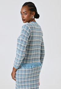 Giacca in tweed a quadri blu e bianchi con dettaglio frangiato all'orlo, caratterizzata da un design aderente e maniche lunghe. Il tessuto testurizzato mostra schemi distintivi.