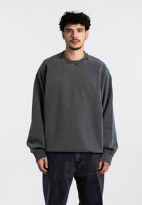Sweatshirt gris de gran tamaño hecho de una tela suave. Cuenta con un escote redondo y un parche con logo en el pecho. Combinado con pantalones gris oscuro.