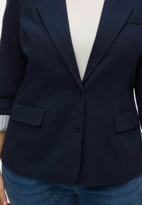 Marineblauwe blazer met een tweeknopsluiting, gekerfde revers en twee voorzakken. De stof is gestructureerd en aansluitend, met een zichtbaar wit binnenvoering.