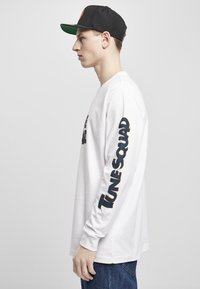 Hvit langermet skjorte med "TUNE SQUAD" trykket i fet svart og oransje på venstre erme. Bæres med en svart caps og blå jeans.