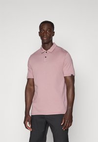 Polo-shirt rose clair en tissu coton avec des manches courtes, patte de boutonnage à trois boutons et un petit logo sur la manche. Porté avec un pantalon sombre.