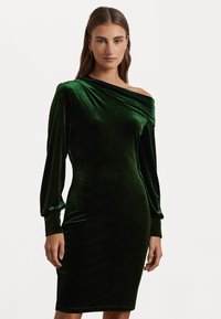 Μακρύ μανίκι πράσινο φόρεμα από βελούδο, με ένα ώμο και ντραπαρισμένο ντεκολτέ. Εφαρμοστό σιλουέτα, μέχρι το γόνατο με λεία υφή.