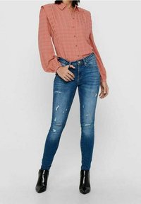Camicia button-up a quadretti rosso chiaro con maniche lunghe, abbinata a jeans skinny blu strappati e stivaletti neri alla caviglia.