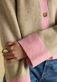 Ruka prekrížená cez béžový a ružový vlnový sveter, na ruke má veľký zlatý prsteň, pričom sú viditeľné modré džínsy v oblasti pásu.