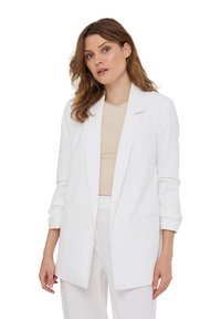 Blazer bianco con una vestibilità sartoriale, maniche lunghe e apertura frontale. Due tasche laterali e una texture di tessuto morbida. Indossato sopra un top beige.