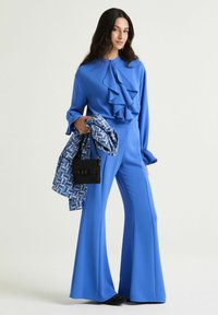 Completo blu brillante con una blusa arricciata e pantaloni a gamba larga. Gli accessori includono una sciarpa con motivo e una piccola borsa nera.
