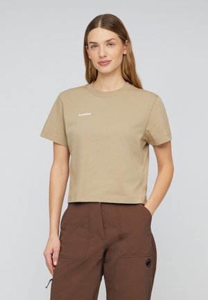 Kvinde med langt lyst brunt hår iført beige t-shirt med korte ærmer og brune bukser med lommer, står foran en ensfarvet lys baggrund.