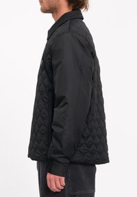 Chaqueta negra acolchada con una capa exterior lisa, cuello alzado y puños de canalé. Cuenta con bolsillos laterales y una silueta aerodinámica.