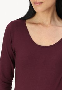 Burgundi hosszú ujjú felső, széles nyakkivágással, puha anyagból készült, sima textúrával és testhezálló fazonnal.