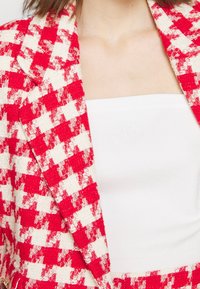 Blazer à carreaux rouges et blancs en tissu texturé, associé à un haut blanc uni sans bretelles avec un logo discret sur la poitrine.