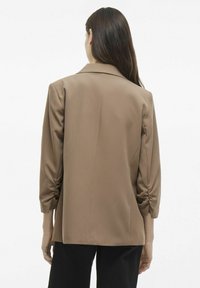 Blazer marrón con un diseño estructurado, hecho de tela suave. Presenta mangas de tres cuartos y sutil fruncido en el puño.