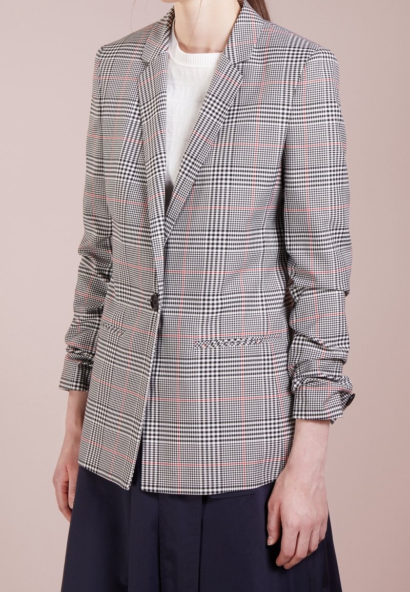 Femme portant un blazer ajusté à carreaux noir et blanc avec des lignes rouges, des manches froncées, un haut blanc et une jupe foncée sur fond beige.