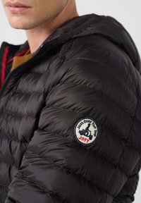 Uomo che indossa una giacca imbottita nera con cappuccio e una toppa rotonda sulla manica raffigurante un motociclista su uno scooter e la scritta "Just Over The Top JOTT".