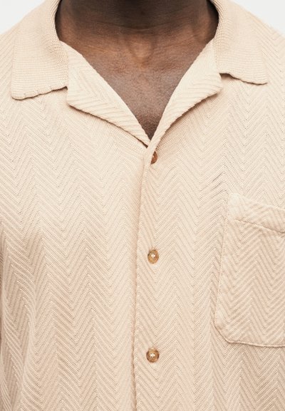 Chemise beige texturée à boutons, avec col et poche poitrine, portée par une personne dont le cou et le haut de la poitrine sont visibles.