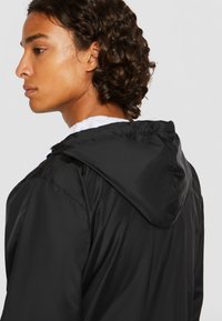 Jachetă cu glugă din nylon negru, cu manșete elastice, având un interior alb. Designul este ușor și aerodinamic, potrivit pentru îmbrăcămintea casual.