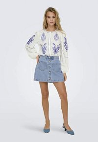 Blusa branca bordada com padrões florais azuis, combinada com uma saia de ganga azul clara com botões na frente e dois bolsos. Sapatos de salto azuis.