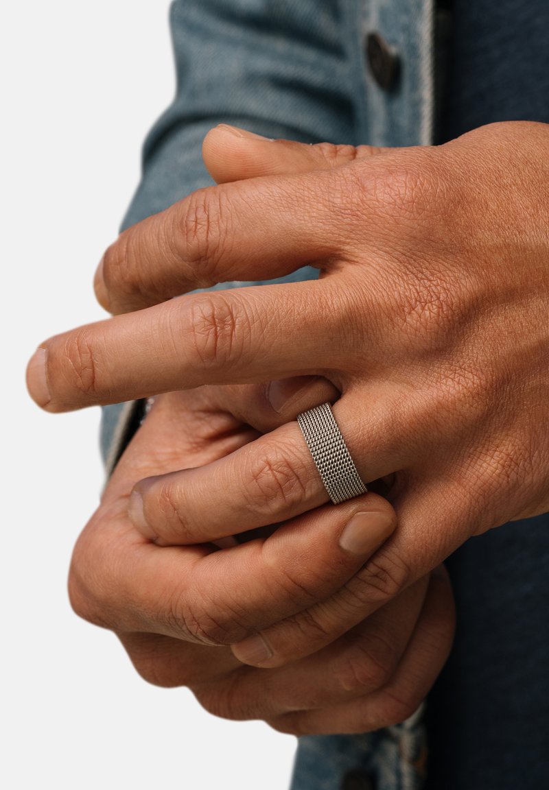 Struktureret sølv ring båret på en hånd med naturlig hudfarve, prydet med en glat overflade og et subtilt netmønster, foran en denim baggrund.