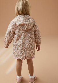 Maluch z blond włosami, ubrany w kwiecistą kurtkę z kapturem, różowe szorty, białe falbaniaste skarpetki i różowe buty, stoi na beżowym tle.