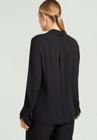 Zwarte blouse met een kraag, lange mouwen met veerdecoraties en een gladde, licht doorschijnende textuur. Achterkant met plooidetail zichtbaar.