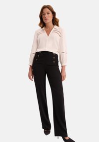 Biała bluzka z przodem na guziki i wycięciami, zestawiona z czarnymi spodniami o wysokim stanie z złotymi akcentami na guzikach i eleganckim krojem.