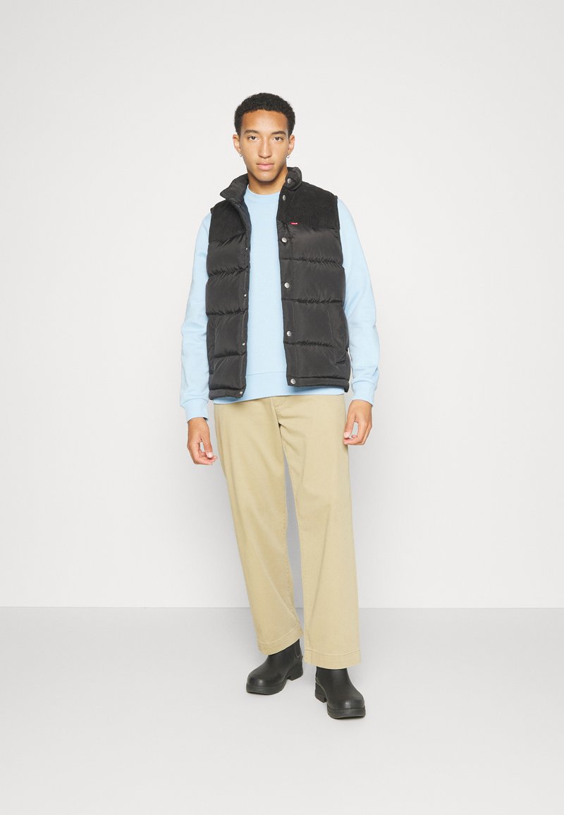 Zwarte puffer vest met een hoge kraag, drukknoopsluitingen en een gestructureerde schouder. Gecombineerd met een lichtblauwe sweatshirt en beige broek.