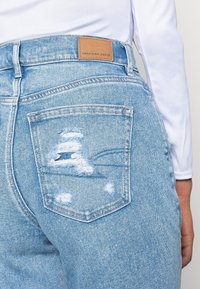Jean en denim bleu clair avec des détails usés sur la poche arrière et une étiquette en cuir beige embossée avec "AMERICAN EAGLE" à la taille.