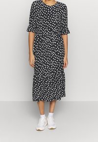 Musta kukallinen mekko valkoisilla auringonliljoilla, lyhyet röyhelöhihoilla ja kerroksellisella helmalla, yhdistettynä valkoisiin lenkkareihin. Puuvillakangasta, polveen ulottuva malli.