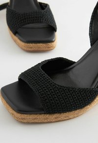 Sandali neri intrecciati con design a punta quadrata, dotati di cinturini texturizzati e tacco a cuneo avvolto in juta. La suola nera liscia valorizza l'aspetto.