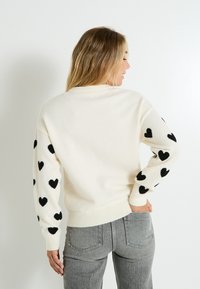 Suéter crema con patrones de corazones negros en las mangas, puños y dobladillo acanalados, y un corte relajado. Combinado con jeans grises.