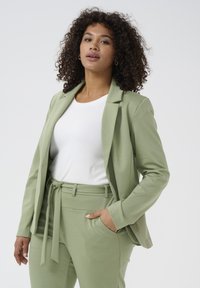 Světl zelený blazer a odpovídající kalhoty z jemné tkaniny. Blazer má klopový límec; kalhoty mají zavázaný pas a kapsy.