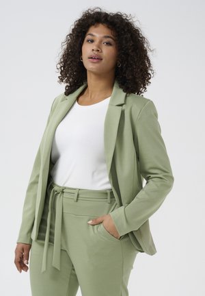 Světl zelený blazer a odpovídající kalhoty z jemné tkaniny. Blazer má klopový límec; kalhoty mají zavázaný pas a kapsy.