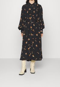 Svart blommig klänning med långa ärmar, krage och bälte i midjan. Har orange och vita blommönster, gjord av ett mjukt tyg.