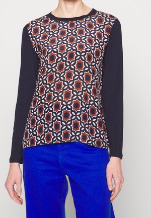 Personne portant un haut à manches longues avec un motif géométrique rouge et bleu à l'avant et des manches noires, associé à un pantalon bleu vif.