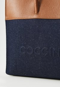 Brown leather and dark blue denim tote bag with embossed logo. Features a textured upper section and smooth lower section.