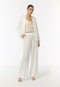 Weißer Blazer über einem beigefarbenen Satin-Top, kombiniert mit weit geschnittenen weißen Hosen und weißen Riemchensandalen; glatte Texturen und taillierte Passform.