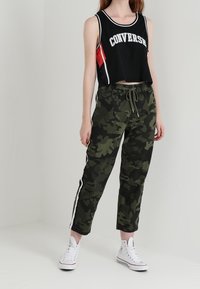 Svart kort t-shirt med "Converse"-bokstäver, tillsammans med gröna kamouflagemönstrade joggers med vita sidostreck och elastisk midja. Vita sneakers.