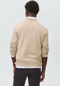 Bež pleteni pulover z dolgimi rokavi, z ribastimi manšetami in robom, nošen čez belo srajco s kratkimi rokavi, viden s hrbta. Gladka tekstura.