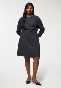 Abito maglione grigio scuro con maniche lunghe, cinte in vita da una cintura sottile. Tessuto strutturato e taglio a ginocchio. Tacchi alti neri a punta.