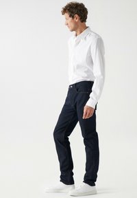 Vaqueros de mezclilla oscura con un corte recto, combinados con una camisa blanca de botones. El modelo está de perfil, usando zapatillas blancas. Diseño sencillo, sin patrones.