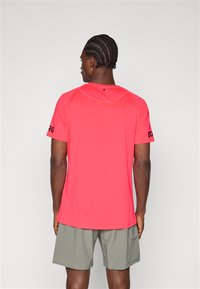 Red athletic t-shirt with short sleeves, smooth texture, round neckline, and black logo accents on sleeves, paired with gray shorts.