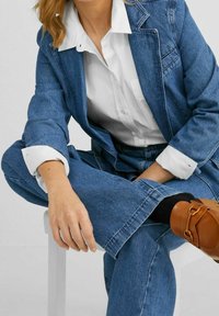 Modrý džínový kostým se strukturovaným sakem a volným střihem kalhot, doplněný bílou košilí a hnědými mokasínami se zlatým detailem.