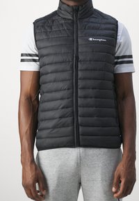 Černá prošívaná vesta s vysokým límcem a předním zipem, se znakem Champion na levé části hrudi. Nositá přes šedé pruhované tričko.