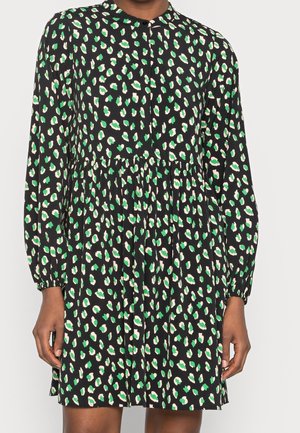 Vestido negro de manga larga con cintura fruncida y un patrón abstracto de manchas verdes y blancas por todo el vestido, llevado por una persona.