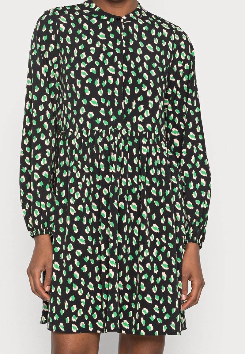 Robe noire à manches longues avec une taille froncée et un motif abstrait tacheté vert et blanc sur l'ensemble, portée par une personne.