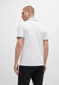 Cămașă polo albă cu guler decorat cu un finisaj închis, mâneci scurte și o textură simplă. Purtați cu pantaloni negri, arătând din spate.