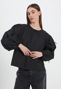 Blusa nera con maniche a sbuffo, scollatura arricciata e dettagli a fiocco sulle maniche. Realizzata in un tessuto morbido, abbinata a jeans scuri.