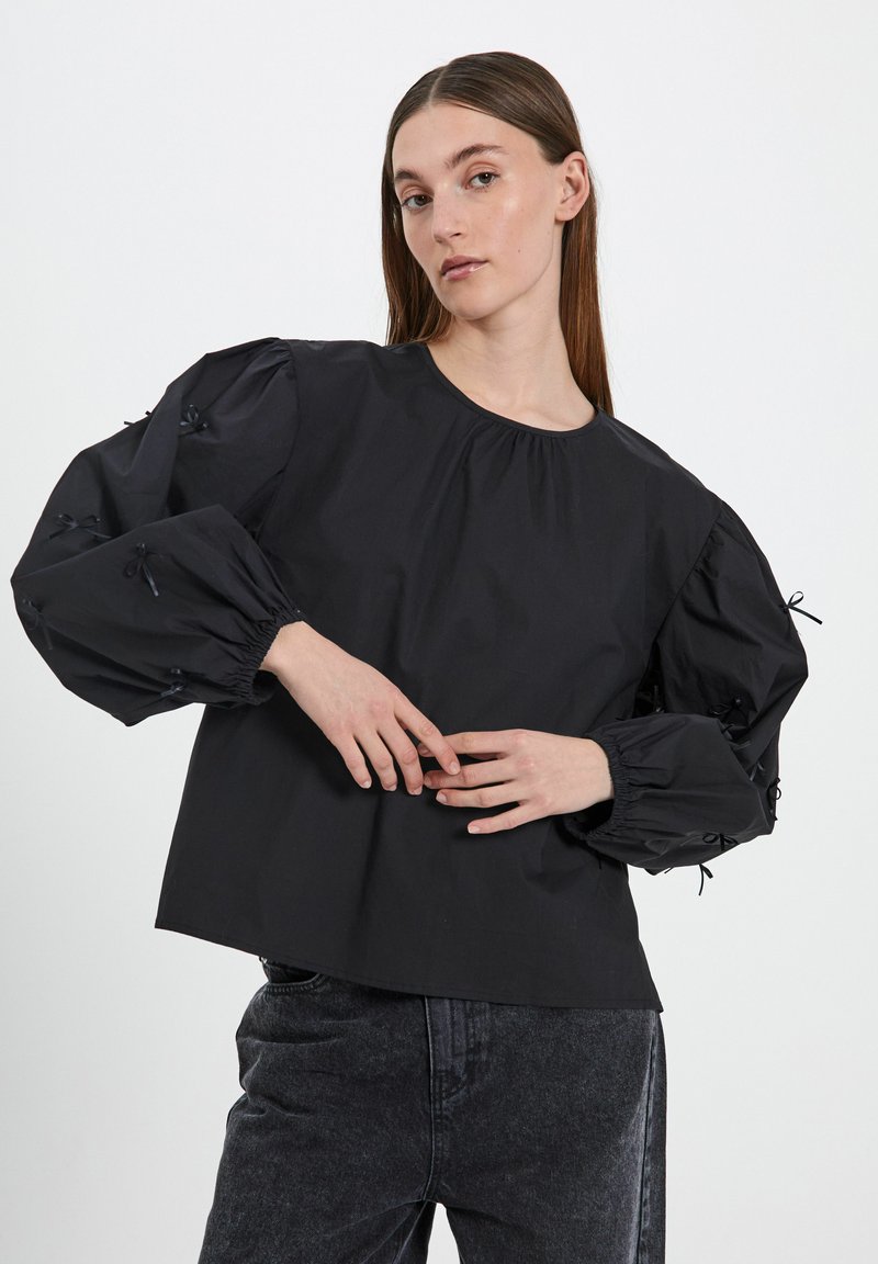 Blusa nera con maniche a sbuffo, scollatura arricciata e dettagli a fiocco sulle maniche. Realizzata in un tessuto morbido, abbinata a jeans scuri.