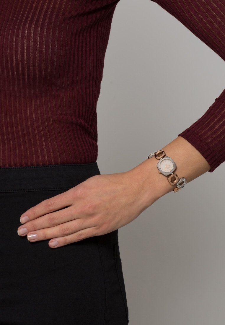 Femme portant un haut bordeaux côtelé à manches longues et une jupe noire, montrant une montre-bracelet en argent et or à son poignet.